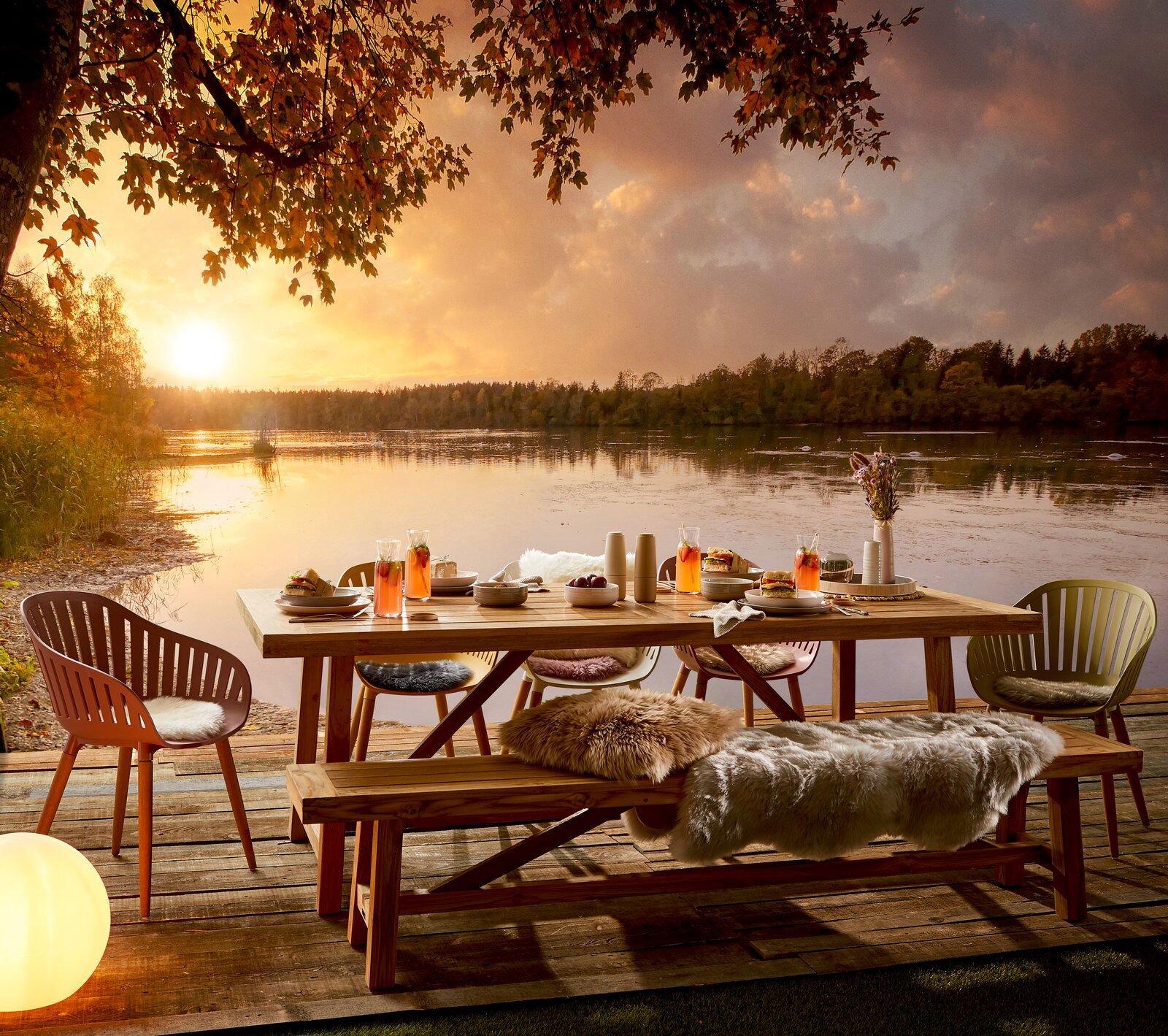 Gartenbank aus Holz auf einer Terrasse mit Blick auf einen See bei Sonnenuntergang, umgeben von Stühlen und einem gedeckten Tisch.