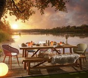 Gartenbank aus Holz auf einer Terrasse mit Blick auf einen See bei Sonnenuntergang, umgeben von Stühlen und einem gedeckten Tisch.