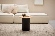 Moderner Beistelltisch mit runder Holzplatte und schwarzem, geripptem Sockel, frontal vor einem cremefarbenen Sofa auf einem Teppich.