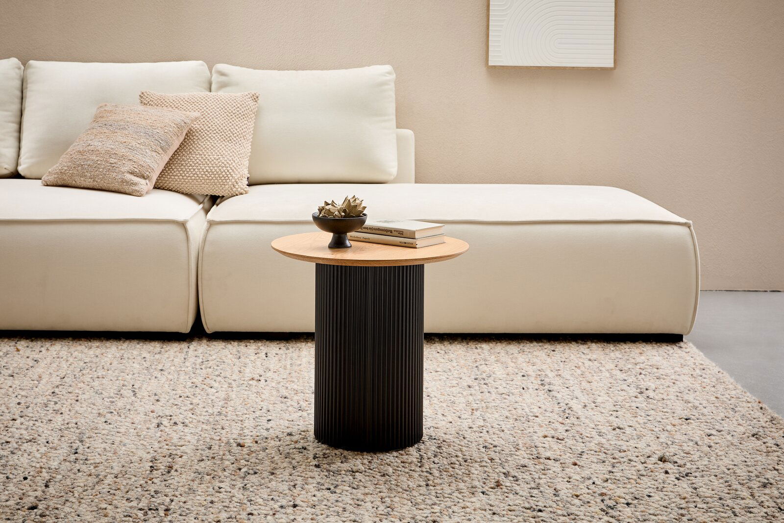 Moderner Beistelltisch mit runder Holzplatte und schwarzem, geripptem Sockel, frontal vor einem cremefarbenen Sofa auf einem Teppich.