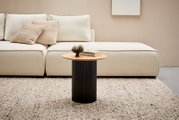 Moderner Beistelltisch mit runder Holzplatte und schwarzem, geripptem Sockel, frontal vor einem cremefarbenen Sofa auf einem Teppich.
