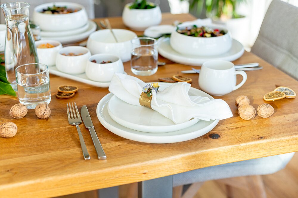 Schlichtes weißes Geschirr-Set auf gedecktem Holztisch mit Tellerstapel, Schalen, Tassen, Gläsern, Besteck und Serviette mit Ring; Perspektive: leicht erhöhte, schräge Frontansicht.