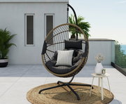 Moderner Hängesessel auf einer Terrasse, seitliche Perspektive, mit Kissen und einem kleinen Beistelltisch mit Vase und Getränk.