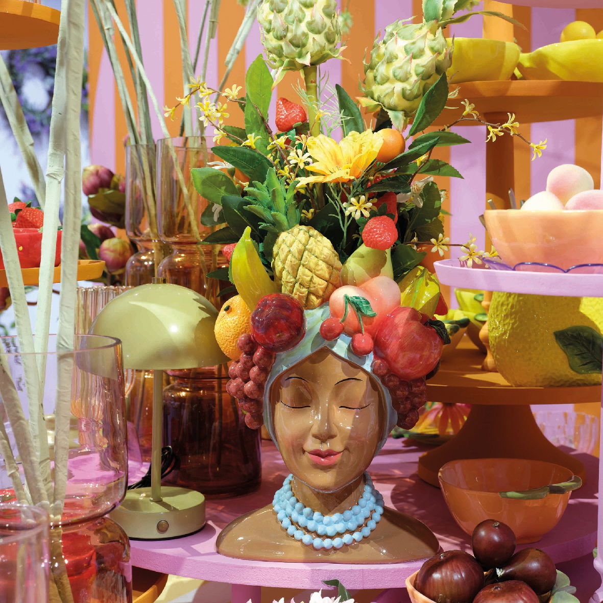 Keramik-Blumentopf in Form eines Frauenkopfes mit buntem Obst- und Blumenarrangement als Kopfschmuck, frontal fotografiert.