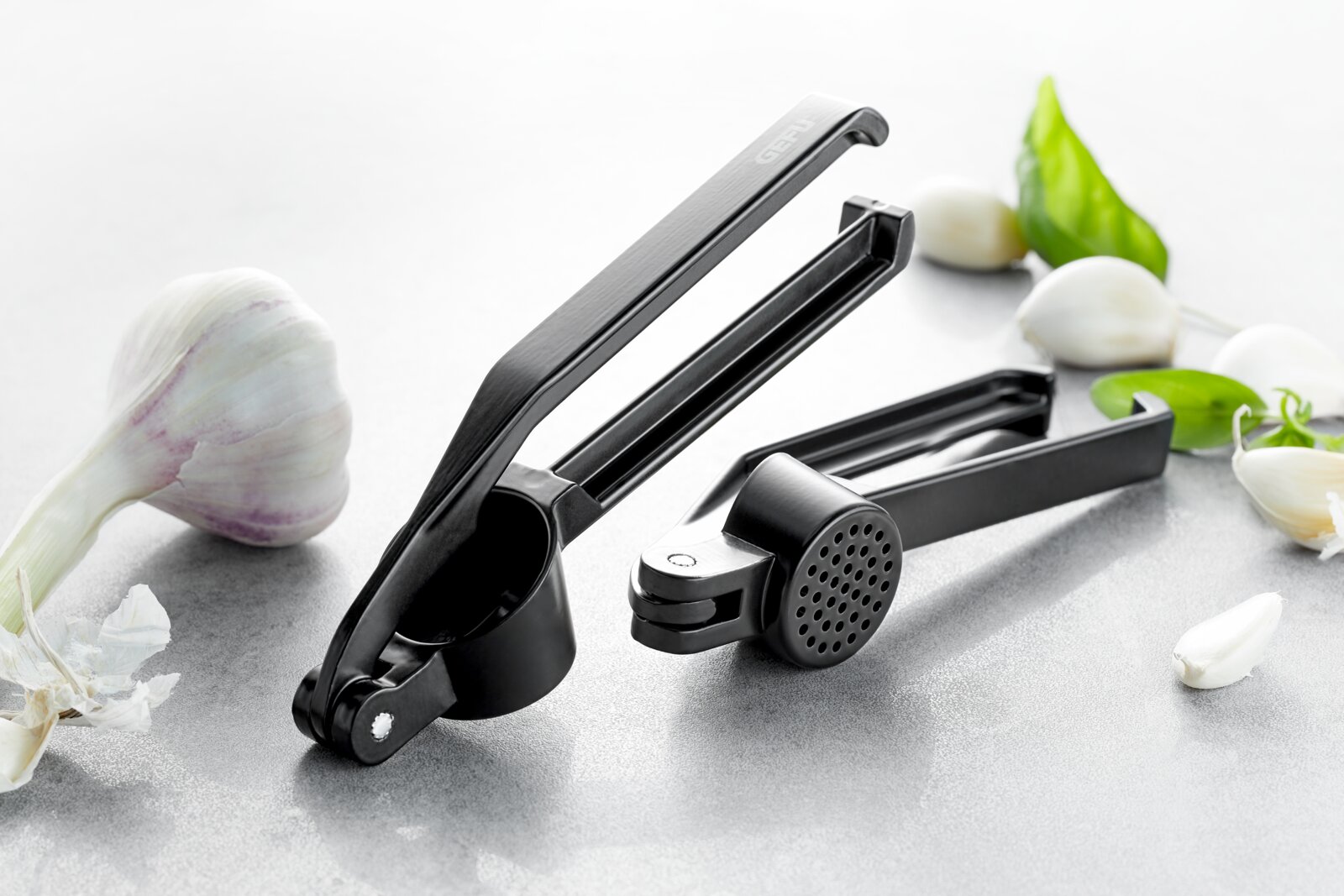 Schwarze Knoblauchpresse aus Metall, geöffnet und in Nahaufnahme auf einer grauen Oberfläche, umgeben von frischem Knoblauch und Basilikumblättern.