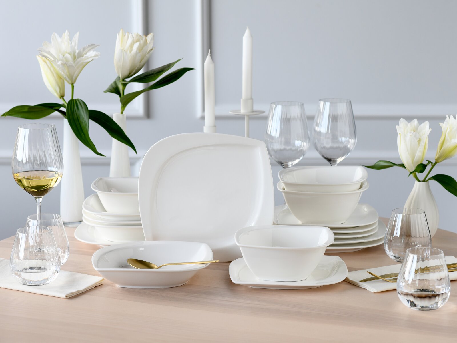 Creatable Tellerset 16-tlg MELBOURNE Modernes weißes Geschirrset mit quadratisch abgerundeten Tellern und Schalen, arrangiert auf einem gedeckten Holztisch mit Gläsern, Kerzen und weißen Blumen, Perspektive: frontale leicht erhöhte Ansicht.