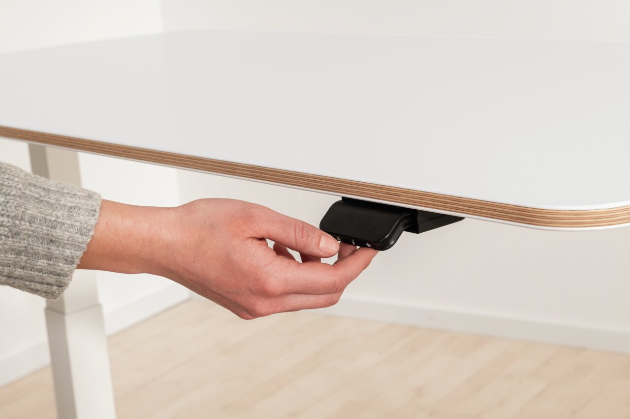 Close-up van een wit bureau met een hand die de hendel voor de hoogteverstelling bedient, gezien vanuit de zijkant.
