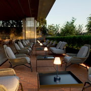 Moderne Tischleuchte Lola Slim 30 auf einem Terrassentisch, umgeben von gemütlichen Sitzgelegenheiten in einer stilvollen Außenumgebung, seitliche Perspektive.