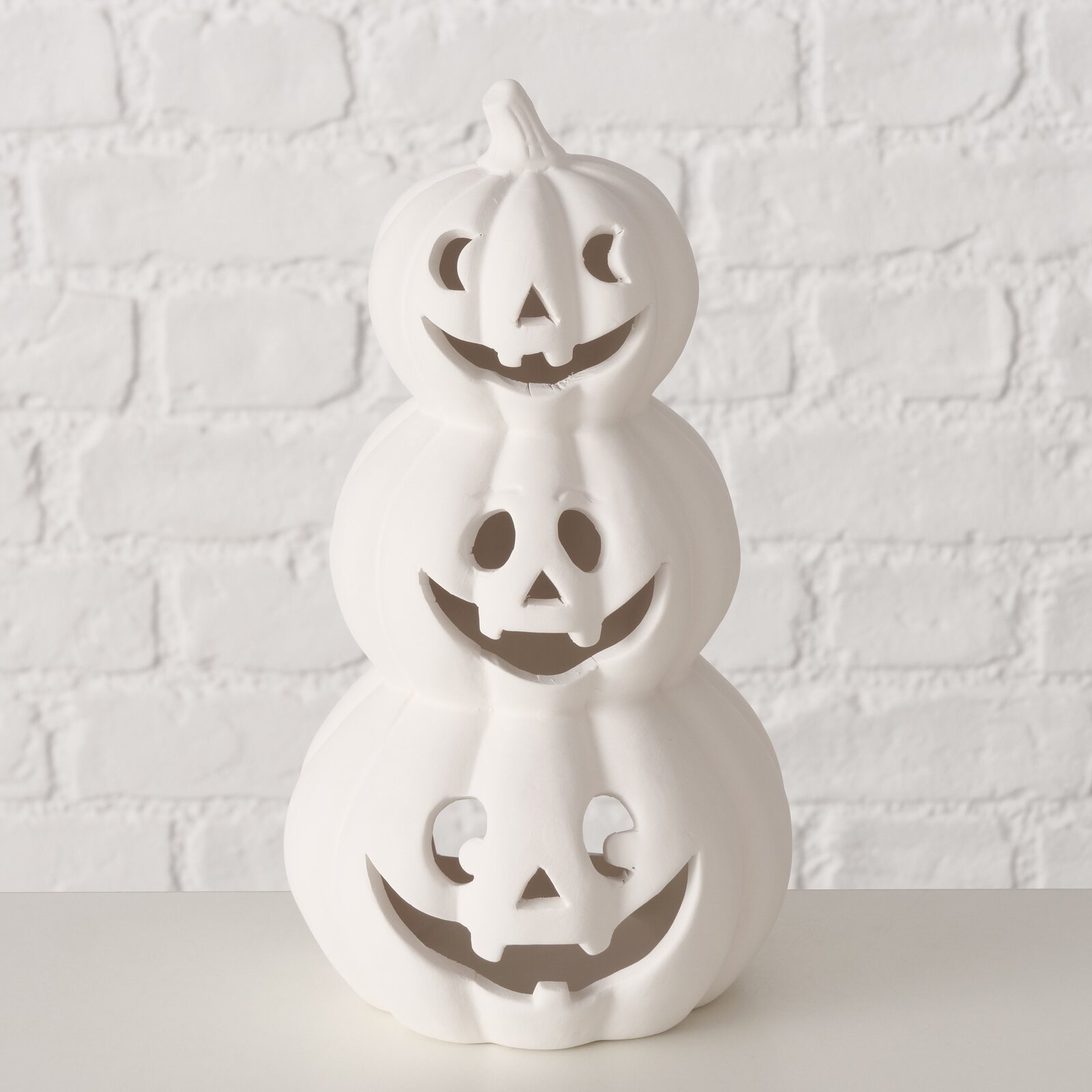 Weißer Teelichthalter in Form von drei gestapelten Kürbissen mit geschnitzten Gesichtern, frontal betrachtet vor einer weißen Ziegelwand.