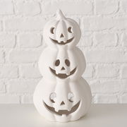 Weißer Teelichthalter in Form von drei gestapelten Kürbissen mit geschnitzten Gesichtern, frontal betrachtet vor einer weißen Ziegelwand.