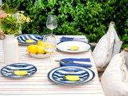 Sommerlich gedeckter Gartentisch mit blau-weiß gestreiftem Geschirr mit Zitronenmotiv, Weingläsern, Zitronenschale und Blumen in weißer Vase auf gestreifter Tischdecke, aufgenommen aus leicht erhöhter Frontperspektive.