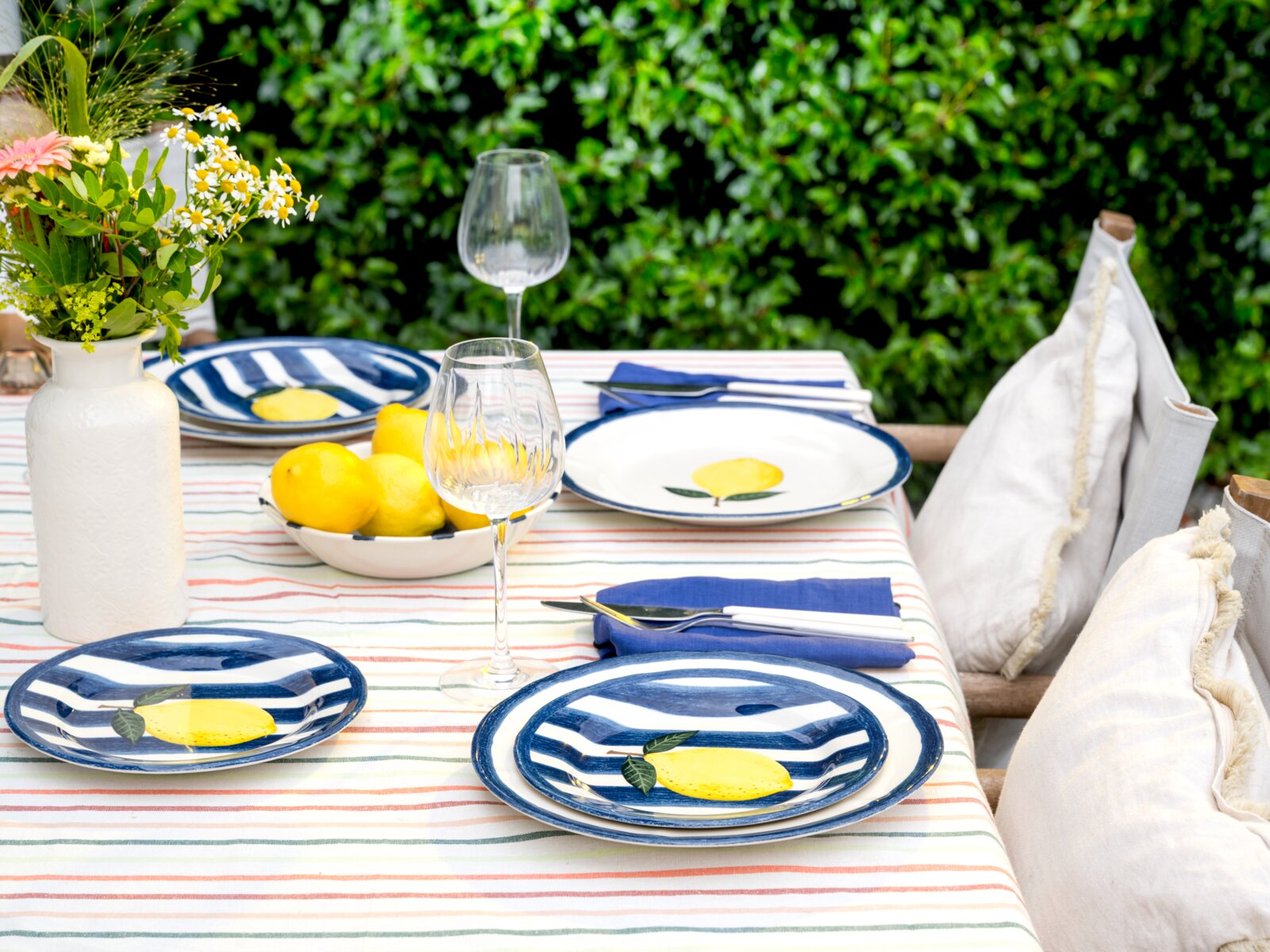 Sommerlich gedeckter Gartentisch mit blau-weiß gestreiftem Geschirr mit Zitronenmotiv, Weingläsern, Zitronenschale und Blumen in weißer Vase auf gestreifter Tischdecke, aufgenommen aus leicht erhöhter Frontperspektive.