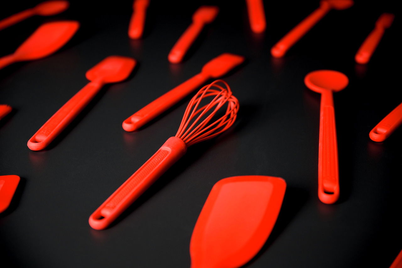 Roter Silikon-Schneebesen in Nahaufnahme, umgeben von weiteren Küchenutensilien auf schwarzem Hintergrund.