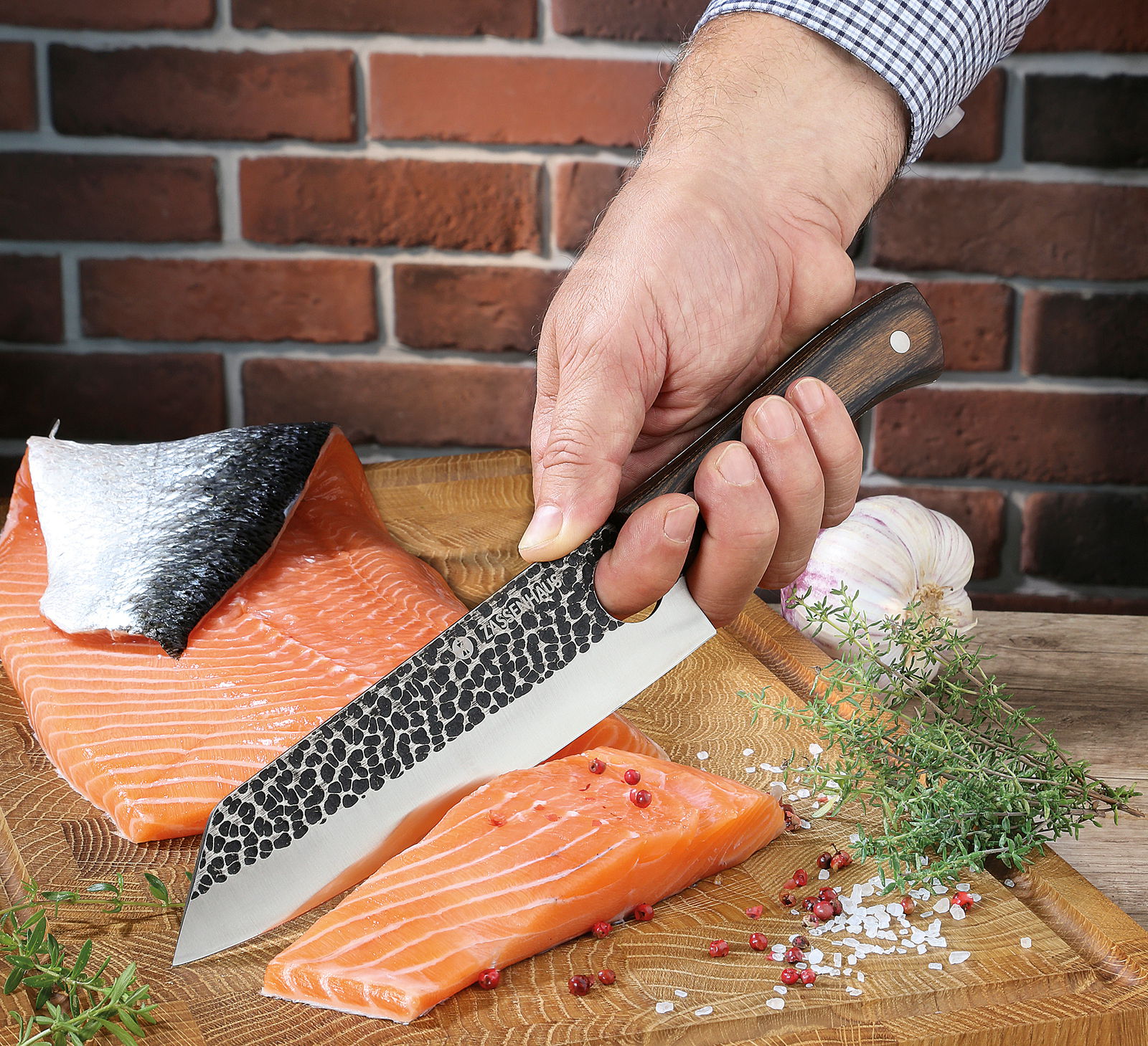 Kochmesser HUNTER 19 cm in der Hand gehalten, schneidet Lachsfilet auf einem Holzbrett, mit Kräutern und Knoblauch im Hintergrund. Perspektive von oben.