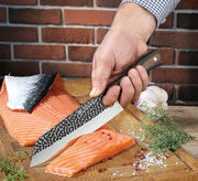 Kochmesser HUNTER 19 cm in der Hand gehalten, schneidet Lachsfilet auf einem Holzbrett, mit Kräutern und Knoblauch im Hintergrund. Perspektive von oben.