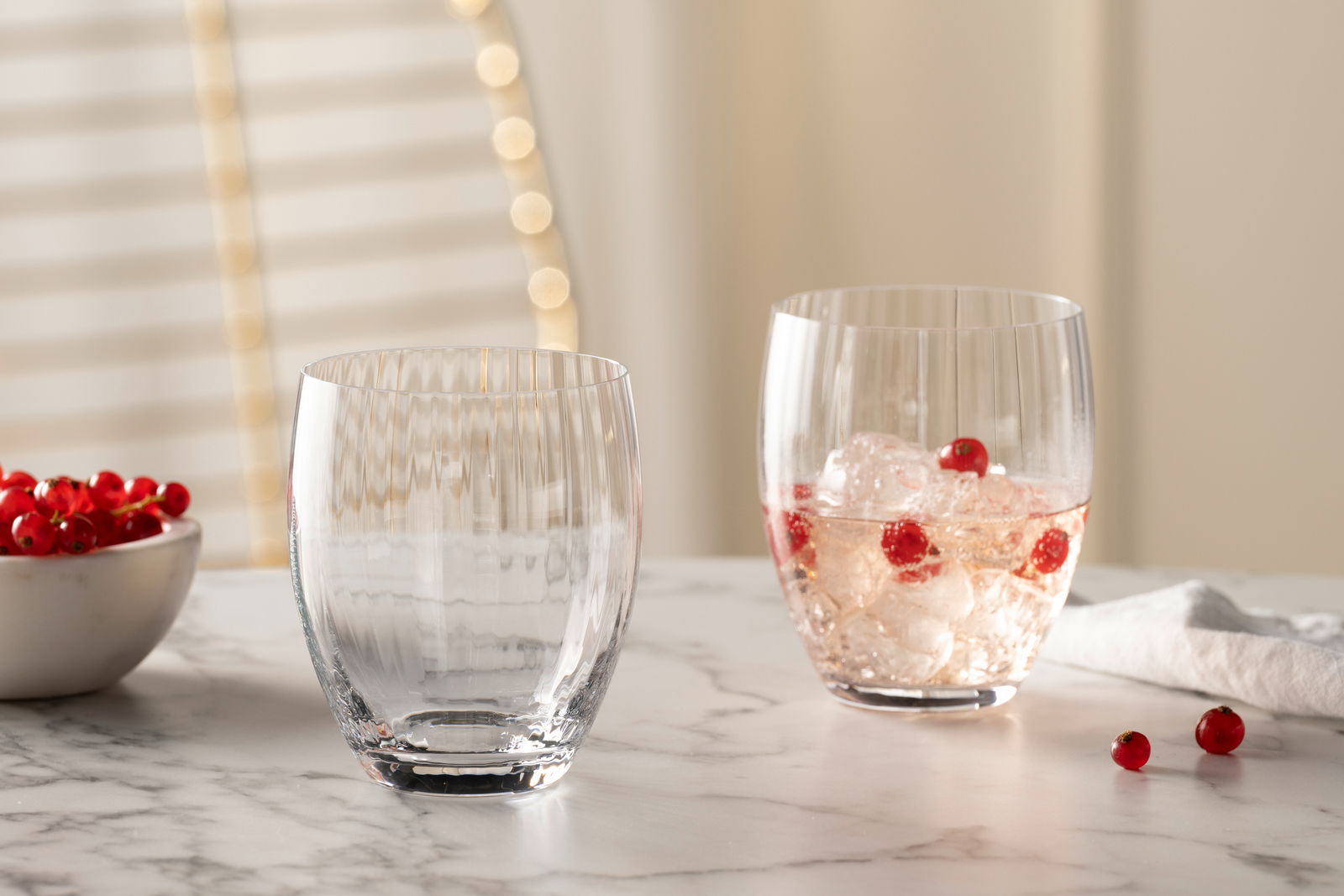 Zwei Trinkgläser POESIA 380ml auf einem Marmortisch, eines leer und eines mit Eiswürfeln und roten Beeren gefüllt, seitliche Perspektive.
