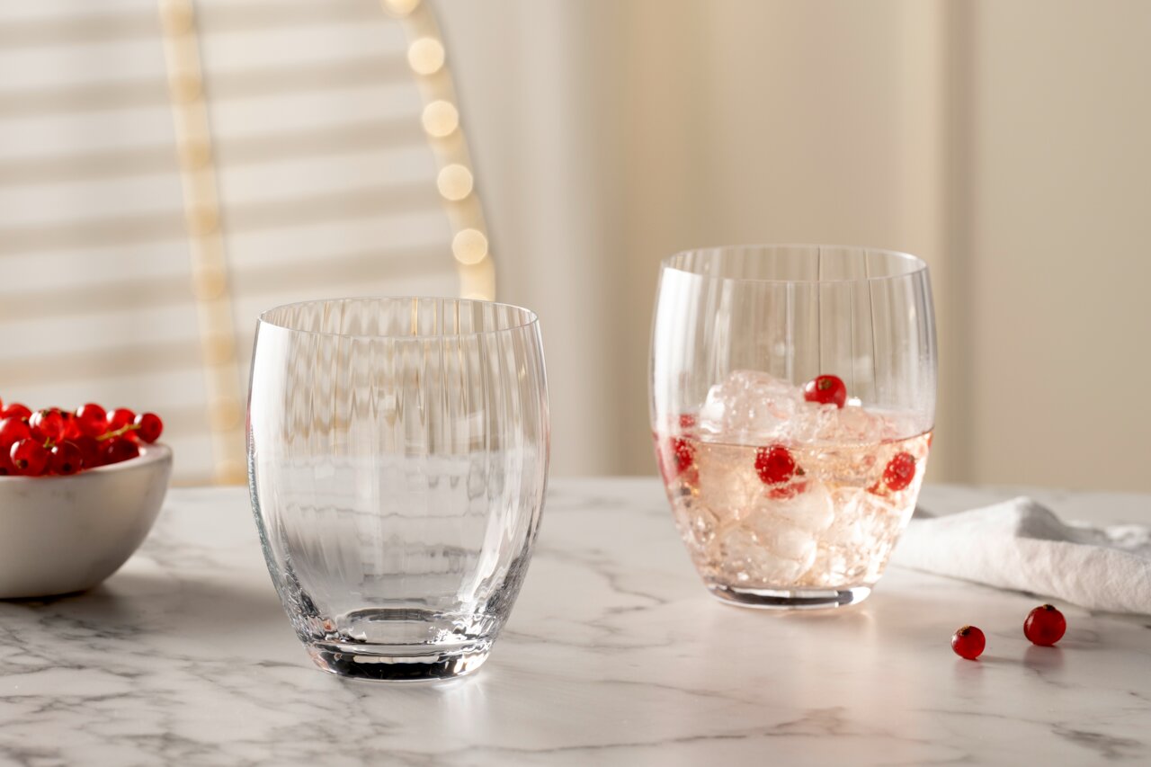 Zwei Trinkgläser POESIA 380ml auf einem Marmortisch, eines leer und eines mit Eiswürfeln und roten Beeren gefüllt, seitliche Perspektive.