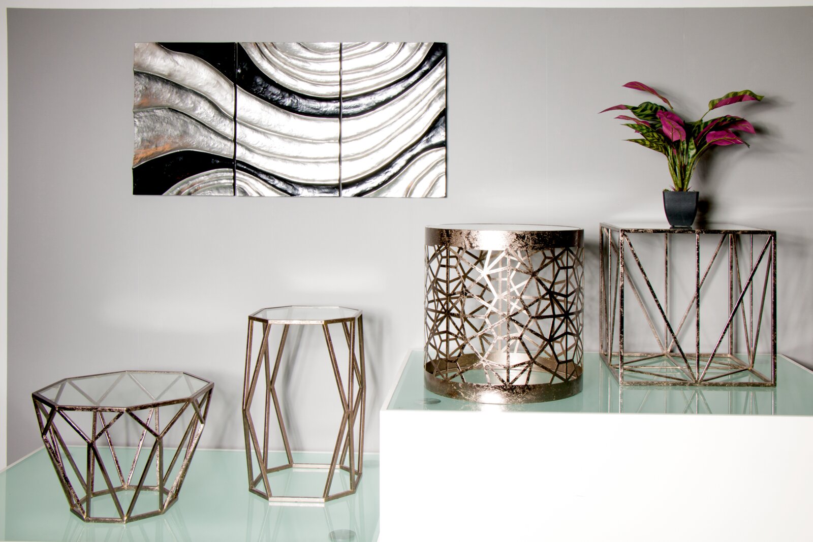 Modernes Wohn-Arrangement in Frontansicht: geometrische Metall- und Glas-Beistelltische/Podeste in Roségold-Optik auf Glasfläche, mit Pflanze und abstraktem Wandbild im Hintergrund