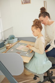 Kind und Erwachsener sitzen an einem Schreibtisch mit einer Schreibtisch-Auflage, die für Bastelarbeiten genutzt wird. Perspektive von der Seite.