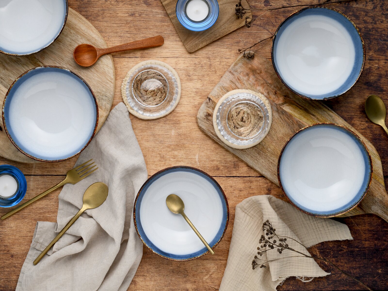 Rustikales Tisch-Arrangement aus der Vogelperspektive: mehrere weiß-blaue Keramikschalen mit goldfarbenem Besteck auf Holzbrettern, Leinentüchern und Holzoberfläche, ergänzt durch Teelichter und Untersetzer.