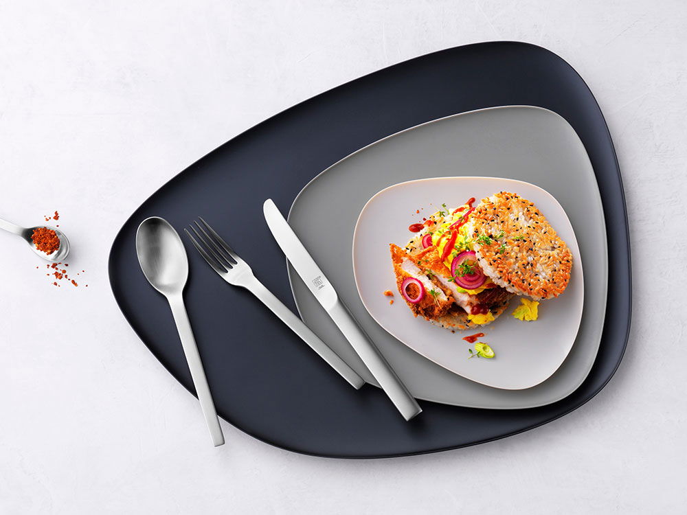 Drei moderne Teller mit Edelstahlbesteck, darauf ein belegter Burger mit Sesam-Brötchen.