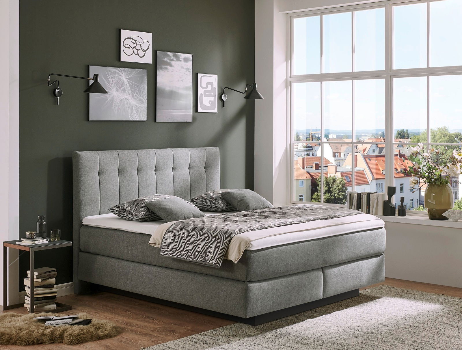 Graues Boxspringbett BX 180/200 in einem stilvoll eingerichteten Schlafzimmer mit grüner Wand und großem Fenster, seitliche Perspektive.