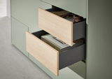Nahaufnahme eines Highboards mit zwei geöffneten Schubladen in hellem Holz und grünem Korpus, gefüllt mit Büchern und Kopfhörern.