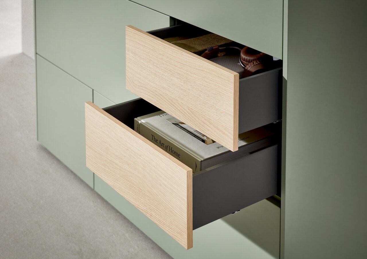 Nahaufnahme eines Highboards mit zwei geöffneten Schubladen in hellem Holz und grünem Korpus, gefüllt mit Büchern und Kopfhörern.