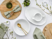 Flatlay/Draufsicht auf ein stilvolles Tischarrangement mit weißer, ornamental geprägter Porzellanschüssel und passender Sauciere, goldfarbenem Besteck, grünen Leinenservietten, Holzplatten, Kerzen und blühenden Zweigen auf hellem Untergrund