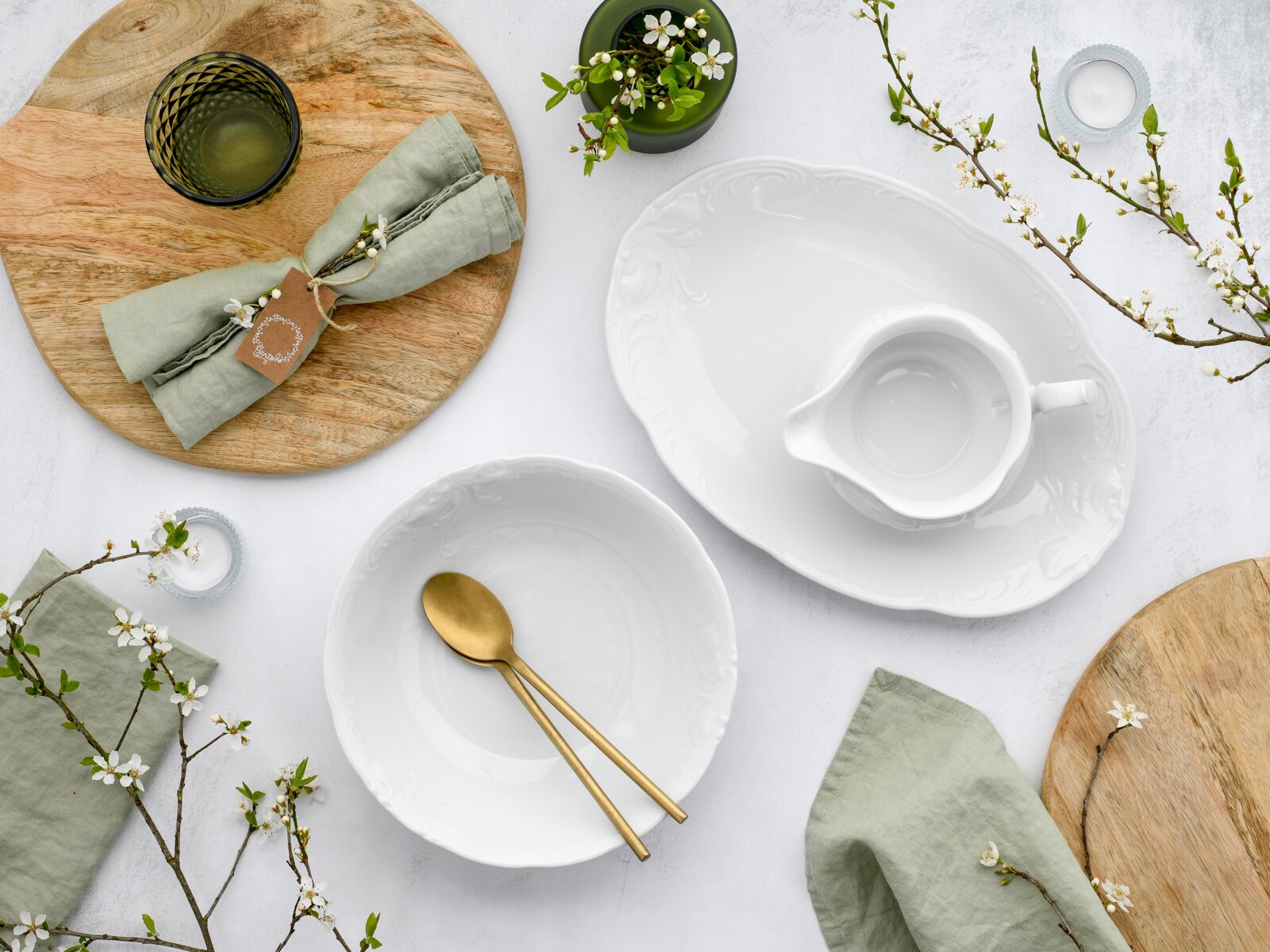 Flatlay/Draufsicht auf ein stilvolles Tischarrangement mit weißer, ornamental geprägter Porzellanschüssel und passender Sauciere, goldfarbenem Besteck, grünen Leinenservietten, Holzplatten, Kerzen und blühenden Zweigen auf hellem Untergrund