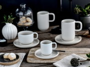 Mattweißes Tassen- und Untertassen-Set in verschiedenen Größen, stilvoll mit Gebäck und Pflanzen auf einem rustikalen Holztisch arrangiert, aus leicht erhöhter frontaler Dreiviertelperspektive.