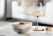 Beige-schwarze Schale ALABASTRO mit Walnüssen, auf einem Tisch neben einem Glas Weißwein und einem Korken, seitliche Perspektive.