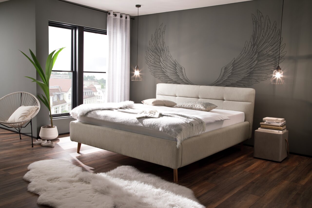 Modernes Polsterbett in einem stilvollen Schlafzimmer mit großen Fenstern, dekorativen Kissen und einer Wand mit Engelsflügeln. Perspektive von der Seite.