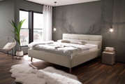 Modernes Polsterbett in einem stilvollen Schlafzimmer mit großen Fenstern, dekorativen Kissen und einer Wand mit Engelsflügeln. Perspektive von der Seite.