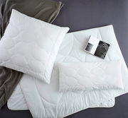 Vogelperspektive auf ein Set aus weißen Kopfkissen und Decken mit kreisförmigem Muster, begleitet von einem grauen Strickstoff und einem geöffneten Buch.