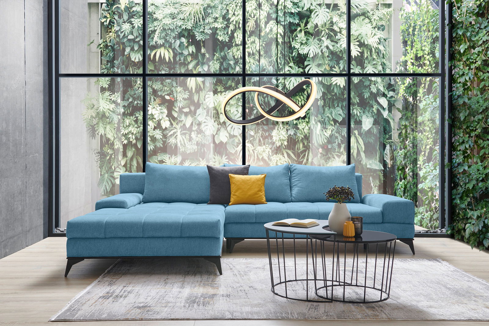 Blaue Eckcouch mit gelbem und grauem Kissen, vor einer großen Fensterfront mit Pflanzenwand, aus der Frontalperspektive fotografiert.