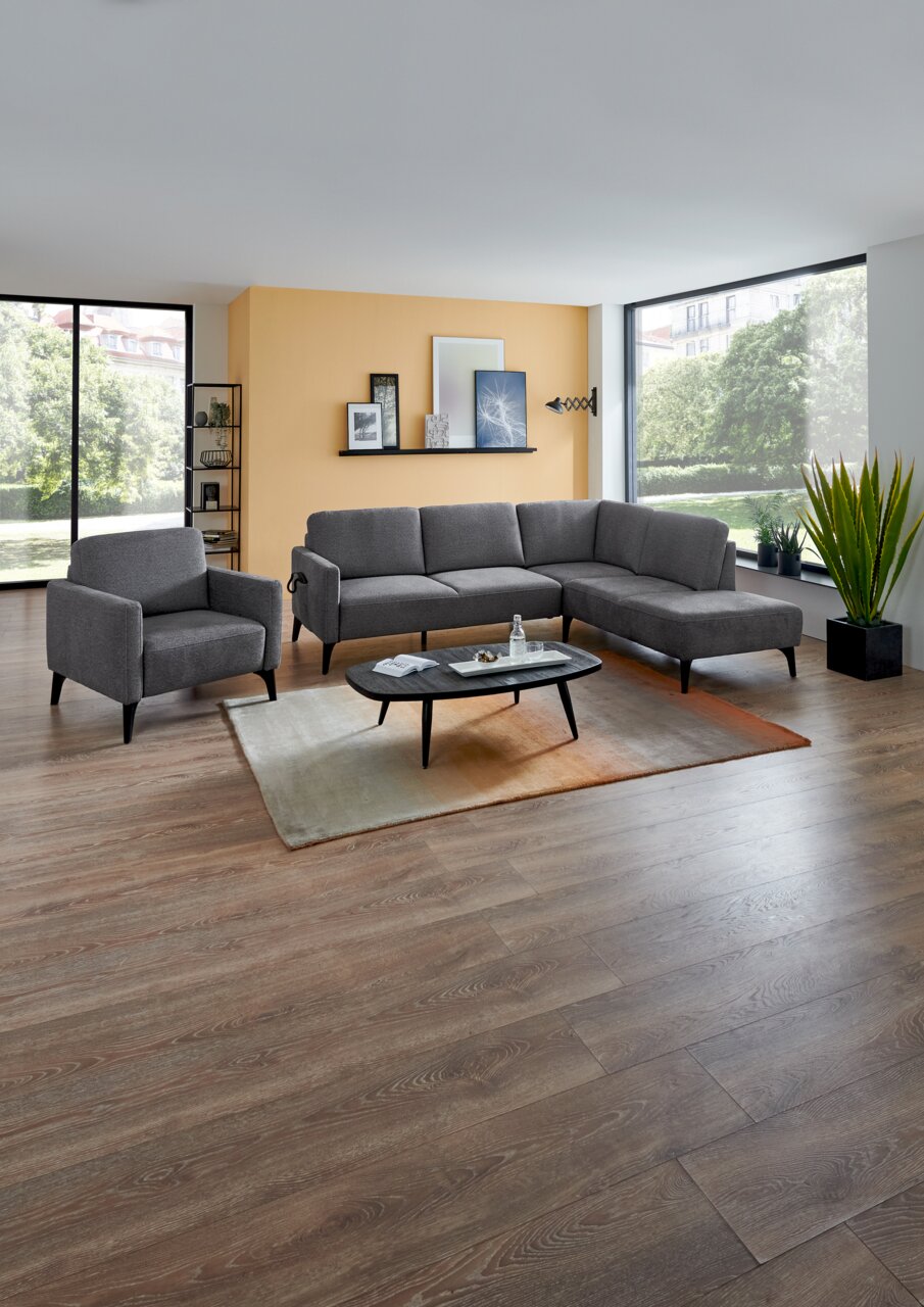 Modernes Wohnzimmer mit grauem Ecksofa und passendem Sessel, Couchtisch auf Teppich vor großer Fensterfront; Perspektive: frontale Weitwinkelansicht auf die Sitzgruppe.