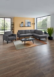 Moderne woonkamer met een grijze hoekbank en bijpassende fauteuil, salontafel op een tapijt voor een grote raampartij; perspectief: frontale groothoekopname van de zithoek.