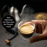 Nahaufnahme eines doppelwandigen Thermo-Espressoglases, das in der Hand gehalten wird, mit einem dampfenden Espresso gefüllt. Im Hintergrund ist eine Kanne zu sehen.