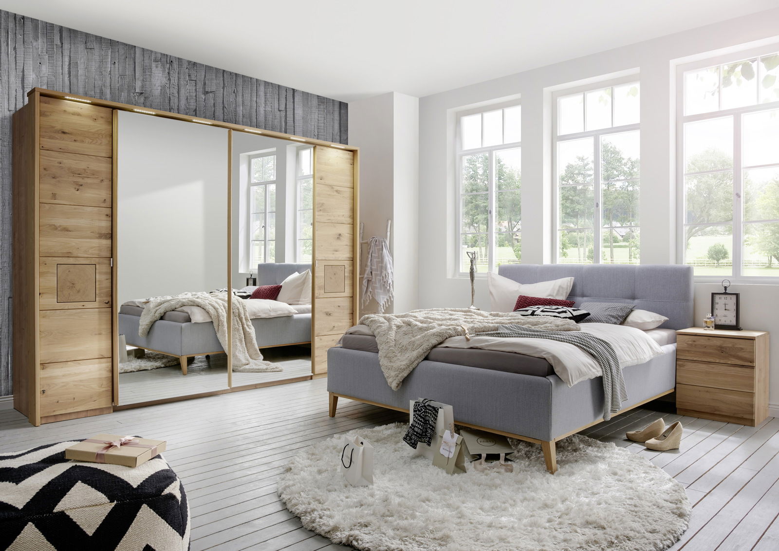 Schlafzimmer mit modernem Schwebetürenschrank aus Holz mit Spiegel in Frontalansicht, grauem Polsterbett und hellem Nachttisch.