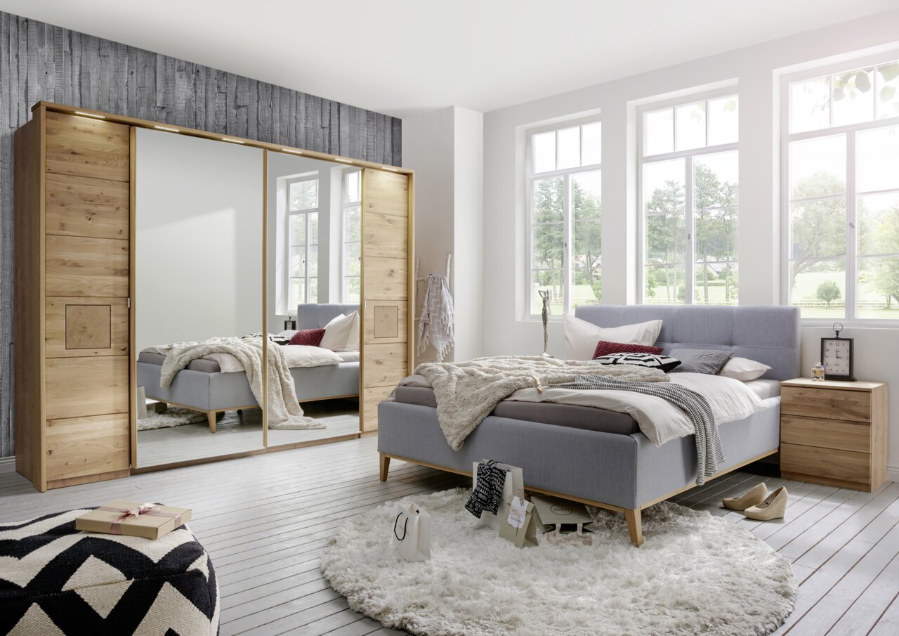 Linea Natura Schwebetürenschrank VITARA Schlafzimmer mit modernem Schwebetürenschrank aus Holz mit Spiegel in Frontalansicht, grauem Polsterbett und hellem Nachttisch.