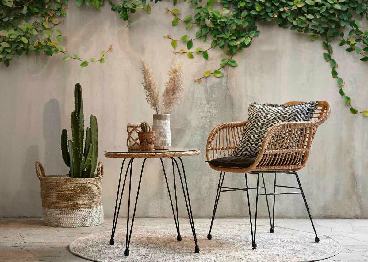 Gartensessel aus Rattan mit schwarzem Kissen, seitliche Perspektive, neben einem runden Tisch und einer Pflanze in einem Korb.