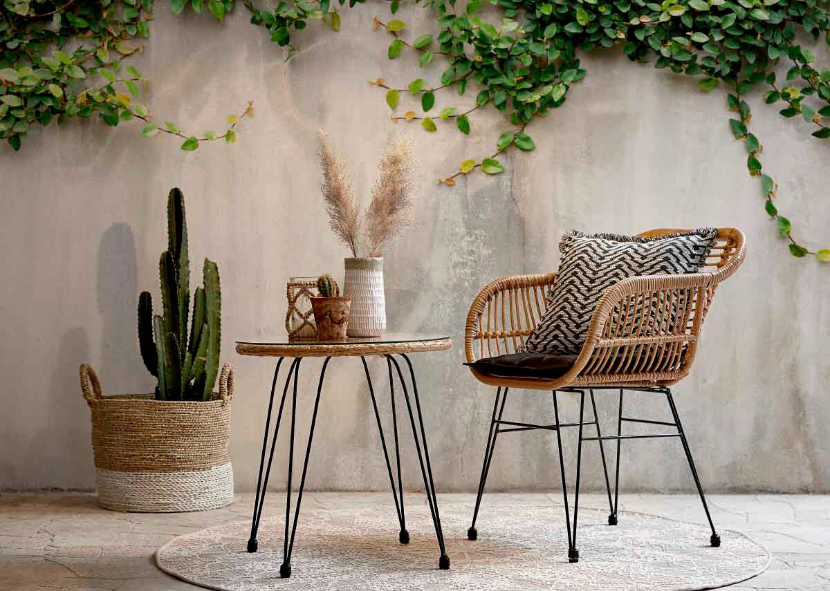 Gartensessel aus Rattan mit schwarzem Kissen, seitliche Perspektive, neben einem runden Tisch und einer Pflanze in einem Korb.