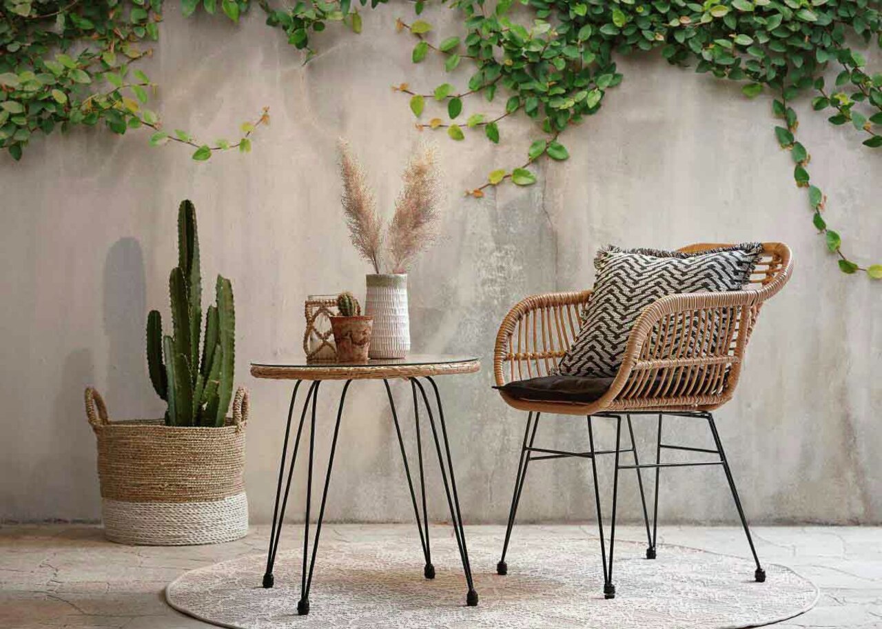 Gartensessel aus Rattan mit schwarzem Kissen, seitliche Perspektive, neben einem runden Tisch und einer Pflanze in einem Korb.