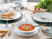 Elegant gedeckter Esstisch mit weißem Geschirr, goldfarbenem Besteck und Glaswaren; im Vordergrund eine Schale Tomatensuppe mit Kräutern aus leicht schräger Perspektive von vorne oben.