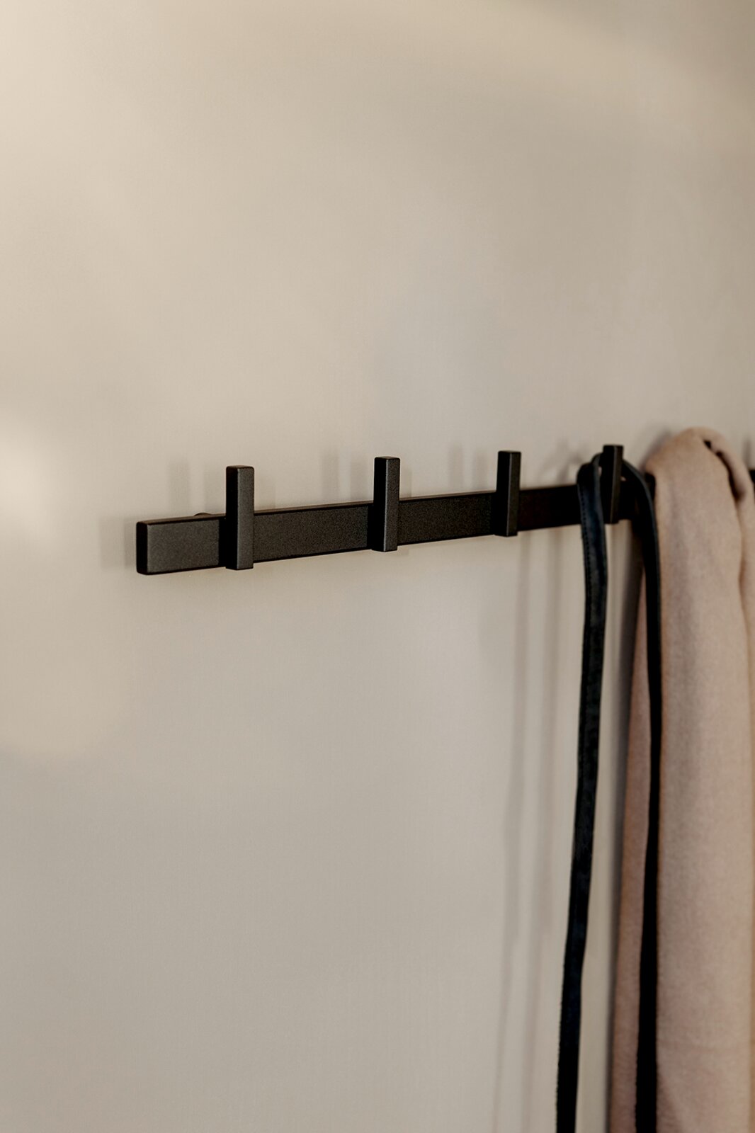 Schwarze, minimalistische Wandgarderobe aus Metall mit vier Haken, seitliche Ansicht an heller Wand, rechts hängen ein Schal und ein Gürtel