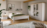 Helles Schlafzimmer mit weißem Holzbett und passendem Kleiderschrank mit Spiegeltüren sowie Nachttisch, aufgenommen aus frontaler Raumansicht leicht von links.