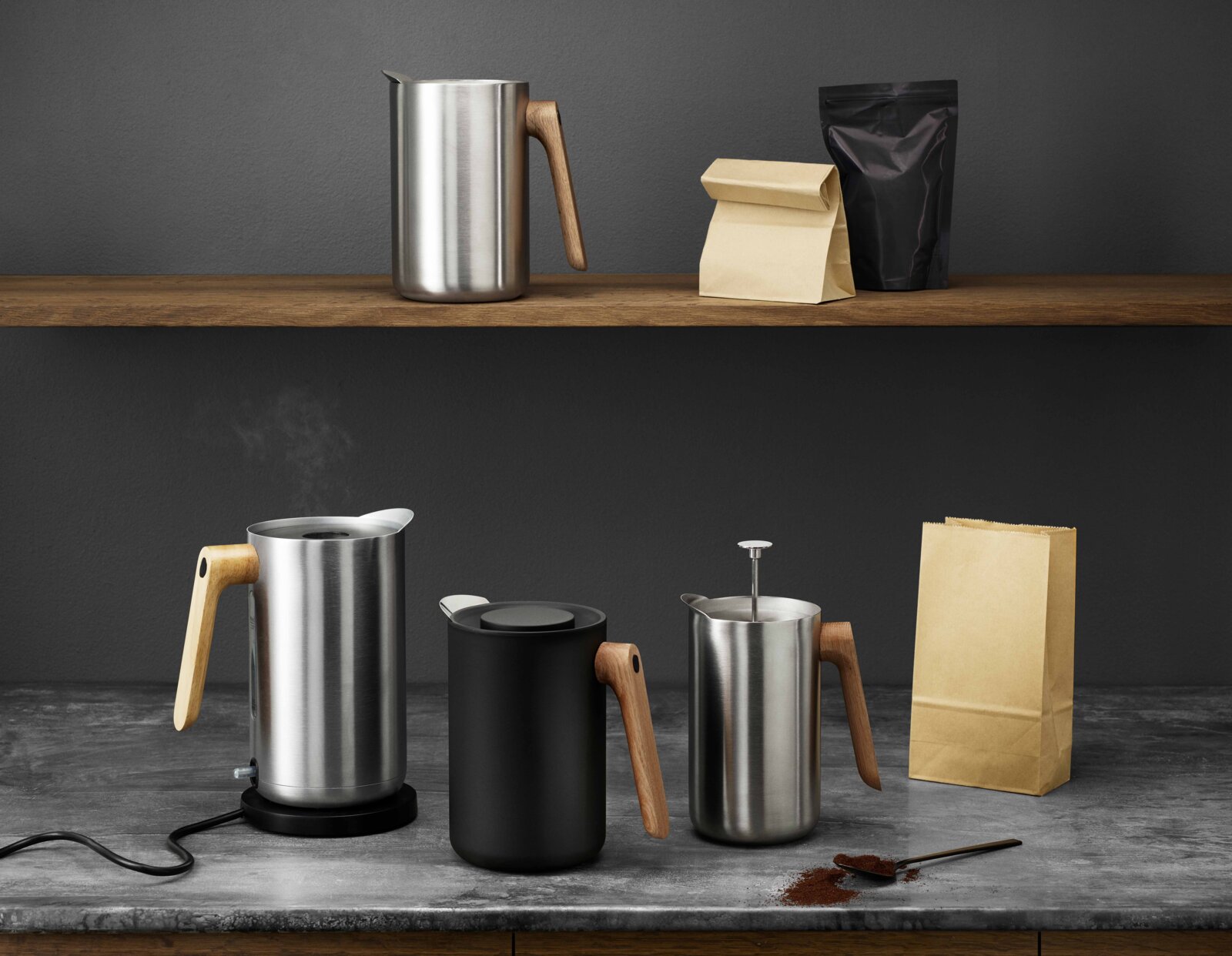 Schuin vooraanzicht van een minimalistische koffieset met roestvrijstalen melkkan en melkopschuimer met houten handgreep op een grijze tafel, met koffiezakken en gemalen koffie op de achtergrond