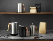 Schuin vooraanzicht van een minimalistische koffieset met roestvrijstalen melkkan en melkopschuimer met houten handgreep op een grijze tafel, met koffiezakken en gemalen koffie op de achtergrond