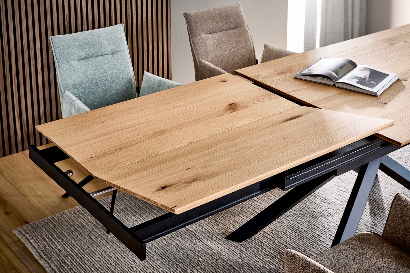 Uitschuifbare eettafel met massief houten blad en zwart metalen onderstel, gedeeltelijk uitgeschoven met zichtbaar uitschuifmechanisme, gefotografeerd vanuit een schuin perspectief van bovenaf in de eethoek.